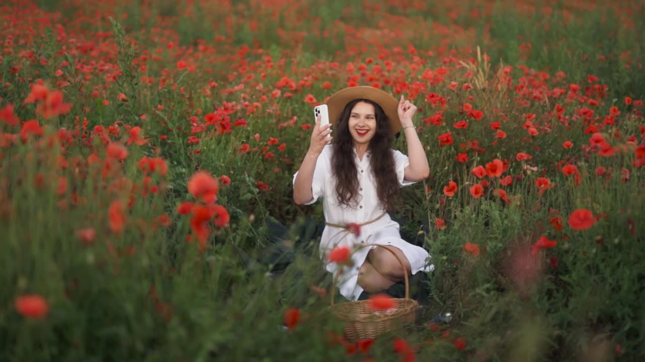 plan general alegre chica de cabello oscuro con un sombrero de paja se sienta en un campo de flores silvestres y amapolas rojas, sonriendo a un mensaje alegre en su teléfono inteligente y soñando