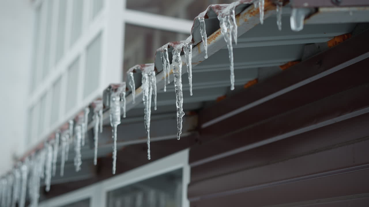 Close-up of metal roof edge covered with long icicles melting gradually, highlighting their intricate frozen structure against a soft blurred background of a building