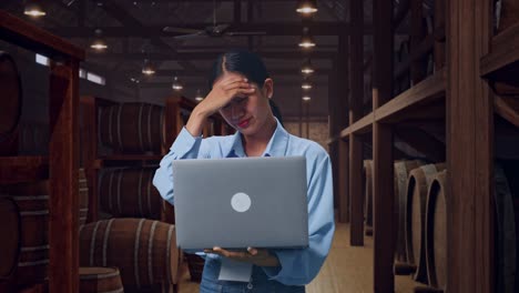asian female professional worker standing with her laptop in cellar of winery, she is nodding her shead with dissapionted