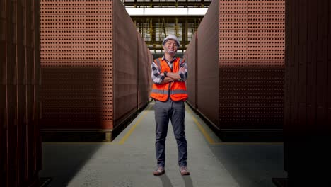 full body of asian male engineer with safety helmet crossing his arms and smiling to camera while standing with red brick packed in stacks are stored