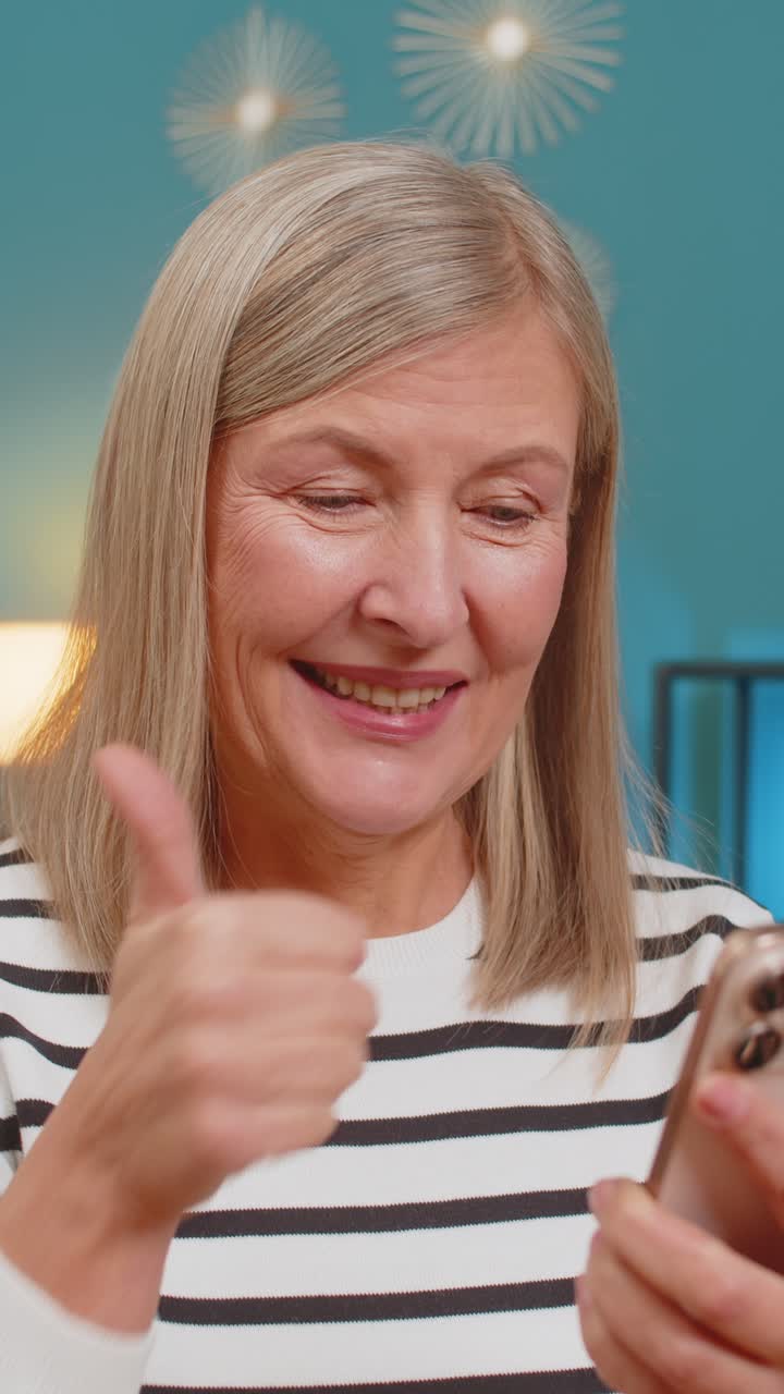Donna matura che guarda le foto dei social media sullo smartphone sorridendo dando il pollice in alto per l'approvazione a casa
