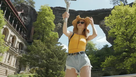 mujer joven en el parque nacional bohemio de suiza, república checa, bajo un arco natural y un viejo hotel grabando video selfie con teléfono inteligente y cardán, cámara lenta