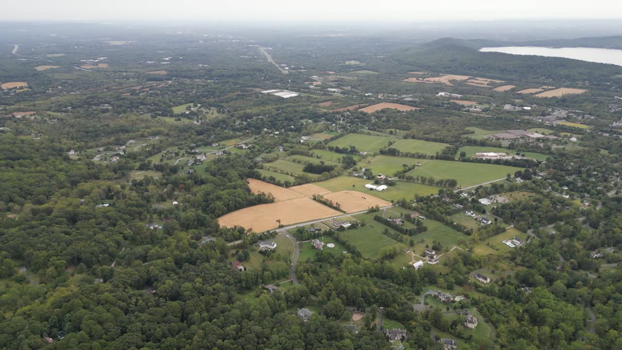 Expansive aerial footage showcasing a rural american landscape, featuring a mix of dense green forests, cultivated farmlands, scattered residential houses, and a distant lake under an overcast sky