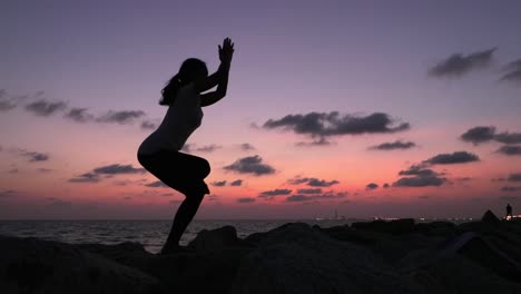 silueta de una mujer practicando yoga en posición de pie con las piernas y los brazos cruzados, disparada durante el crepúsculo cerca del océano