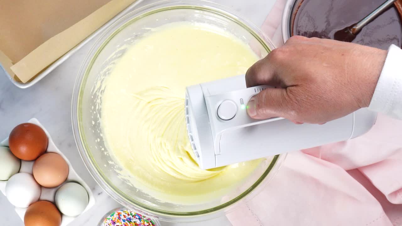 Baking chocolate brownies and using a white electric mixer to mix the batter. Making brownies on a white background surrounded by eggs, pink napkin and a baking pan. In the kitchen making a dessert