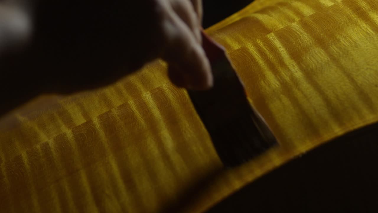 Liutaio applica la vernice alla tavola posteriore di un violoncello, esaltando in tempo reale le fiammature naturali e le venature del legno usando un pennello a mano sotto illuminazione direzionale per profondità e calore.