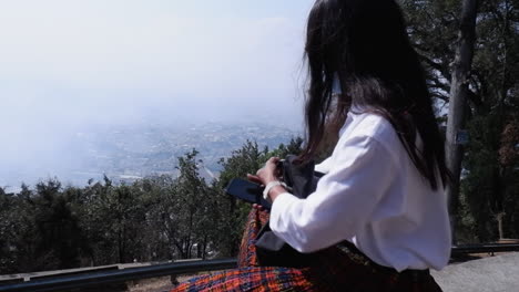 raven haired woman in pleated skirt sits at viewpoint above foggy city