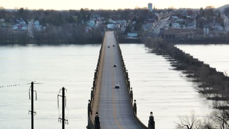 Drone-view-of-vehicles-driving-on-a-two-lane-bridge-leading-to-a-downtown-district-during-sunset-in-the-spring