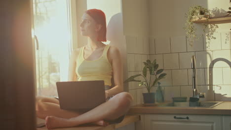 girl sitting on windowsill in sunlit kitchen and working on laptop