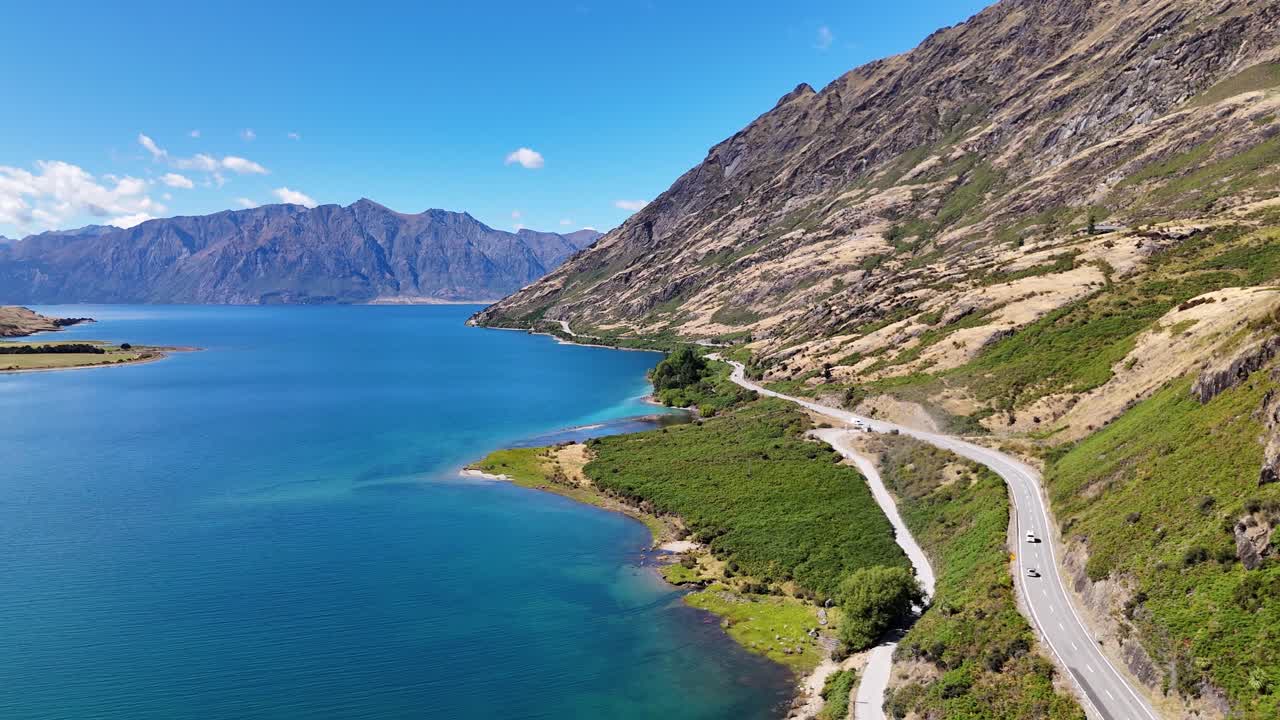 4k drone video of the winding scenic road around beautiful blue Lake Hawea in Wanaka, New Zealand. The lake is surrounded by mountains and green landscapes