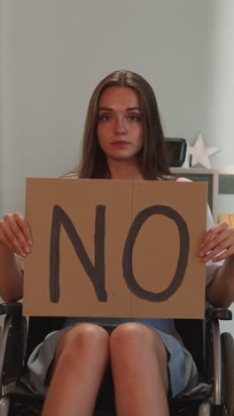 serious woman with disability shows poster with word no to social inequality looking in camera. young person with amyotrophic lateral sclerosis sits in wheelchair