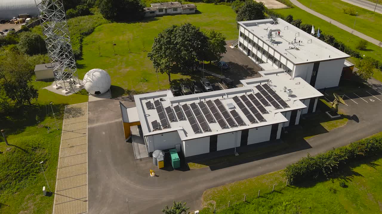 Aerial drone orbiting and flying around a Estonian weather station that has solar renewable energy panels on the roof top collecting energy and radome radar dome is visible next to the building, sunny