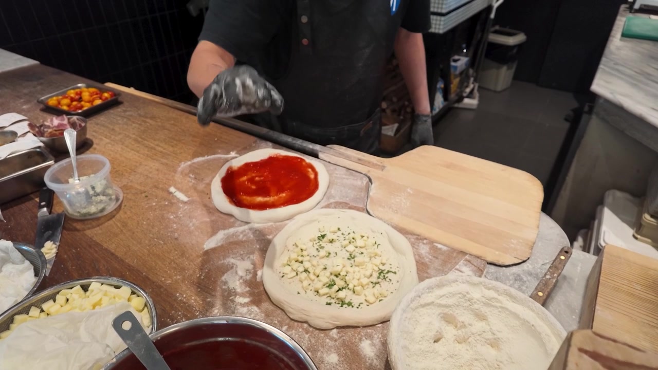 Chef con guanti neri spalma salsa di pomodoro sull'impasto in un ambiente di cucina commerciale.