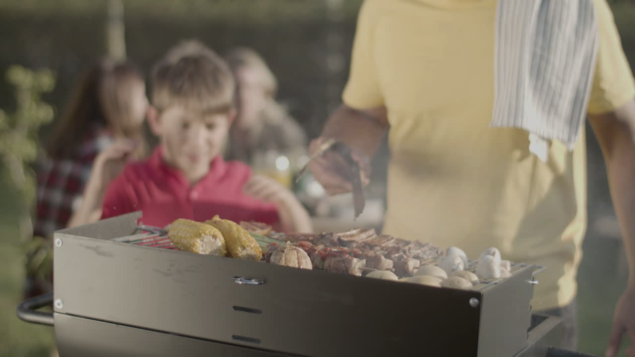 Uomo che gira la carne sulla griglia del barbecue, suo figlio che soffia sul fuoco.