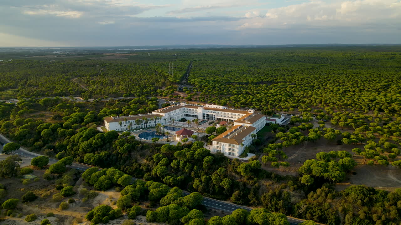 Aerea - Drone che si avvicina lentamente al lussuoso Garden Playanatural Hotel circondato da una fitta foresta di pini marittimi sempreverdi al tramonto, El Rompido, skyline dell'Andalusia, Spagna.