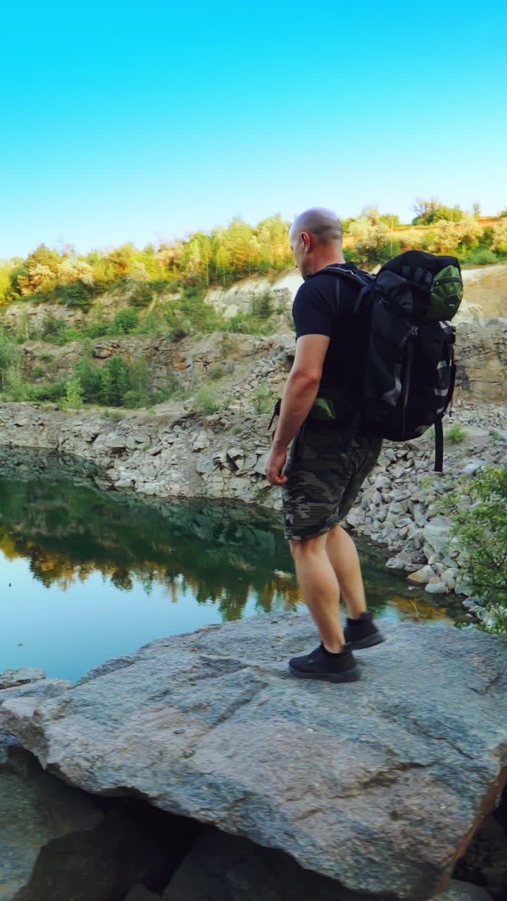 A tourist in summer clothes walks on the edge of a quarry with huge stones and looks at the beautiful lake and the nature around it. Beautiful view. Vertical video