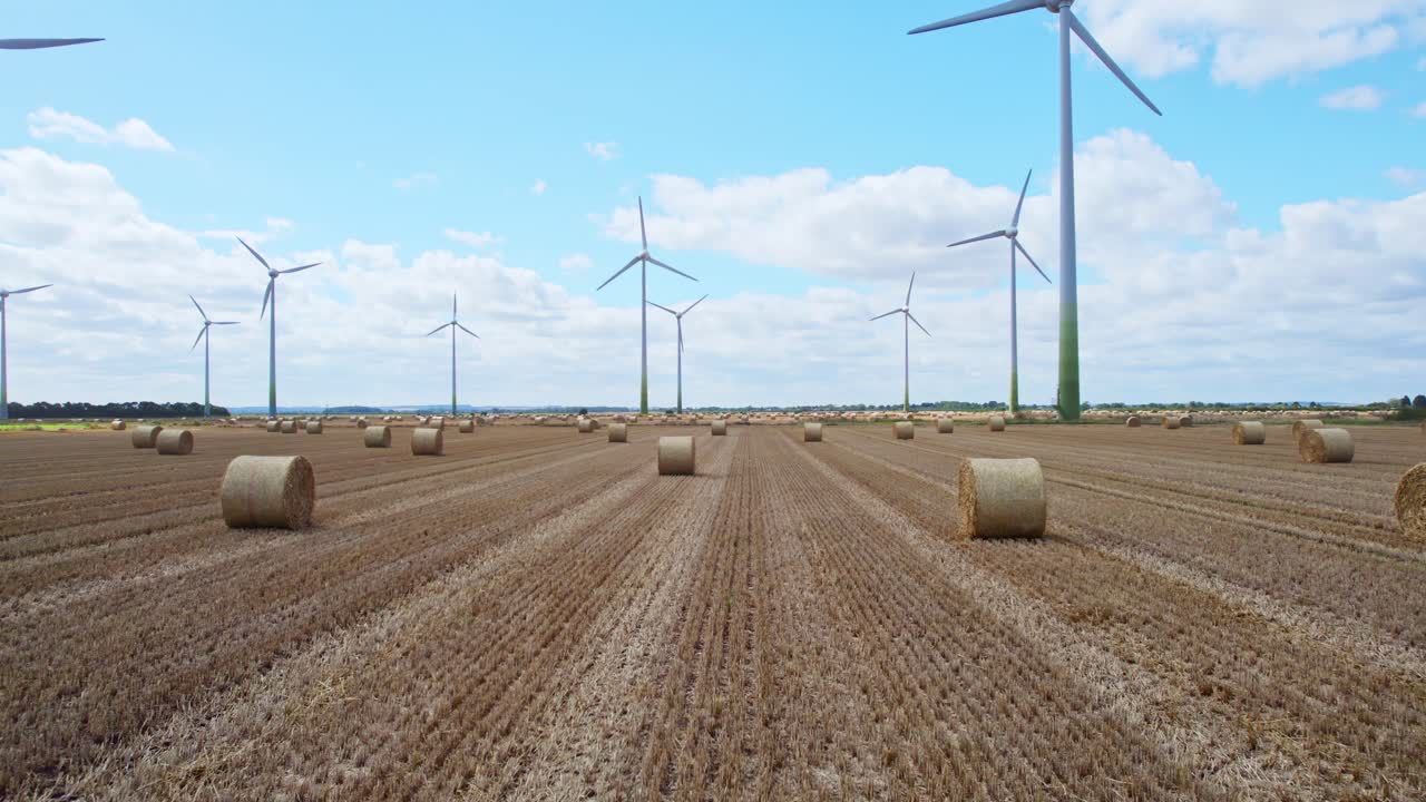 High in the sky, a captivating tableau unfolds as wind turbines spin within a Lincolnshire farmer's newly harvested field, where golden hay bales complete the picture