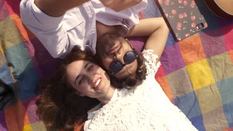 happy lovely young couple lying on colorful blanket in the park taking selfies smartphone romantic with guitar sunglasses beautiful attractive girl top view rotating camera slow motion