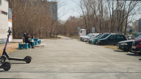 vista cercana de un estacionamiento urbano con un scooter eléctrico en primer plano, coches estacionados y personas relajándose en bancos
