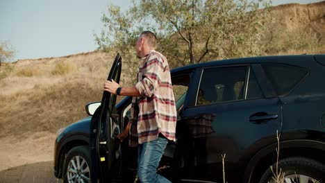 a happy man with gray hair in a plaid shirt gets out of his black one in the car and, together with his girlfriend and daughter, takes out things for a picnic in a steppe area with dry grass in summer