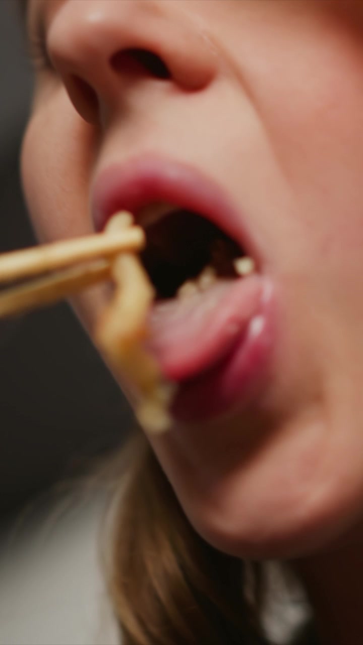 Primo piano di una donna che mangia cibo con le bacchette. La donna tiene gli utensili e li porta alla bocca, concentrandosi sui tratti del viso come labbra, naso e occhi. L'illuminazione soffusa evidenzia la texture della pelle.