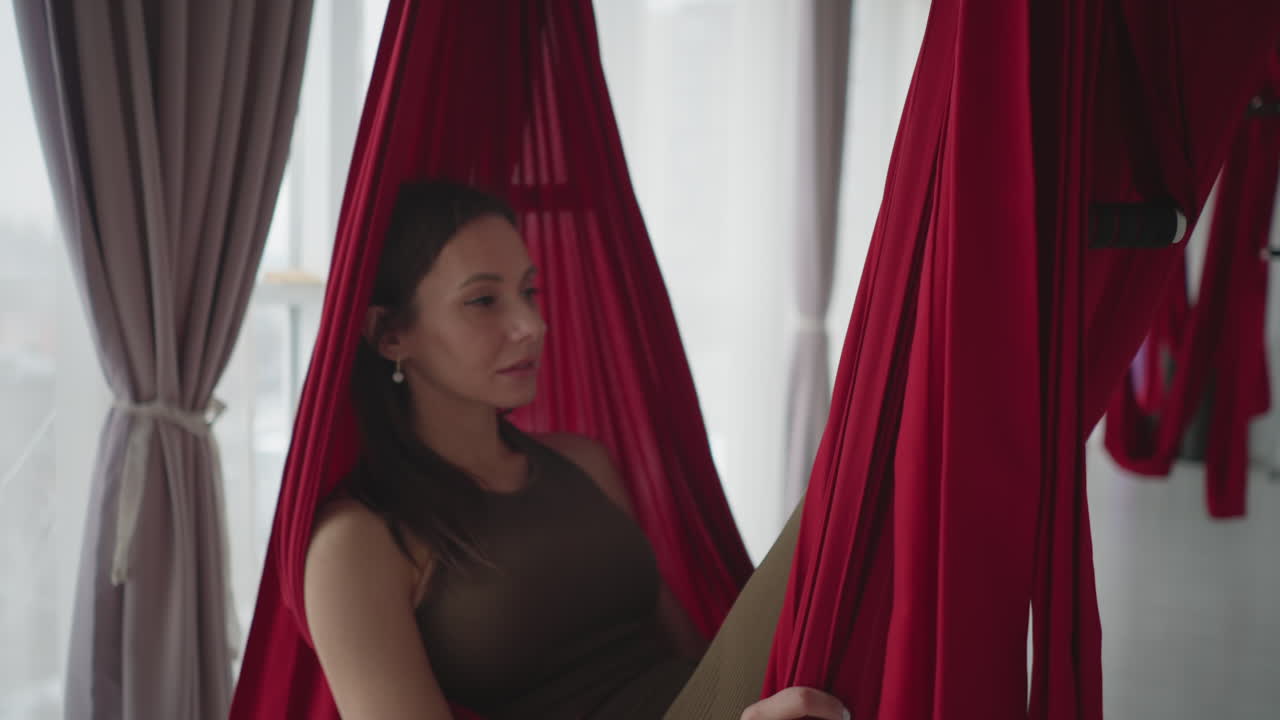 Close up view of female athlete lying thoughtfully in red silk hammock swinging gently in bright studio with sheer curtains and urban window view conveying balanced tranquil calm reflection