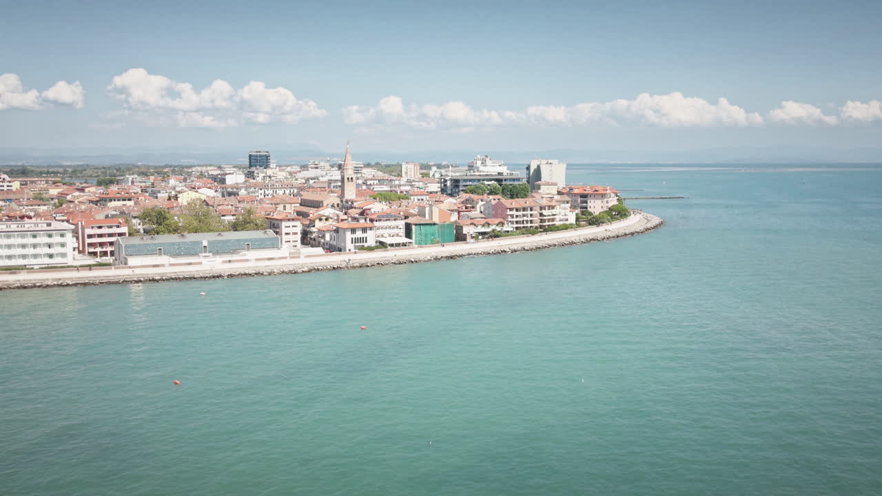 Ripresa aerea cinematografica in 4K UHD con drone in avvicinamento sopra il blu del Mare Adriatico verso la storica costa dell'isola di Grado. Ideale per promozioni turistiche italiane, pubblicità di vacanze estive e sfondi web.