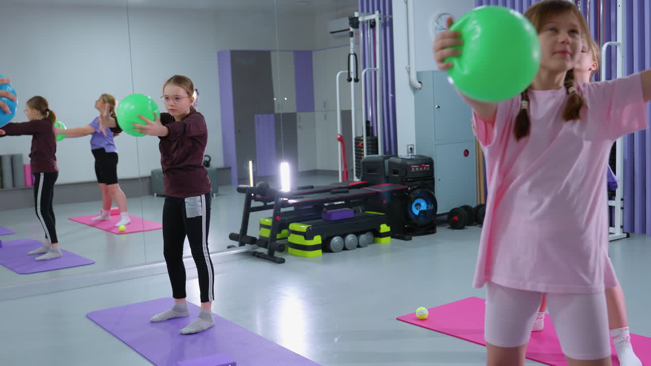 los niños realizan ejercicios de flexibilidad del brazo con globos verdes, equipos de gimnasia y cajas de música visibles en el fondo de un moderno centro de fitness