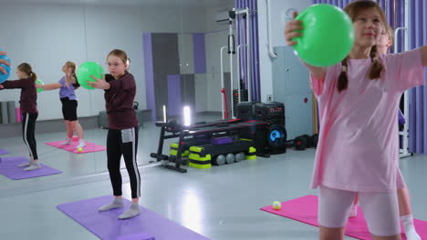 children perform arm flexibility exercise with green balloons, gym equipment and music box visible in background of modern fitness center