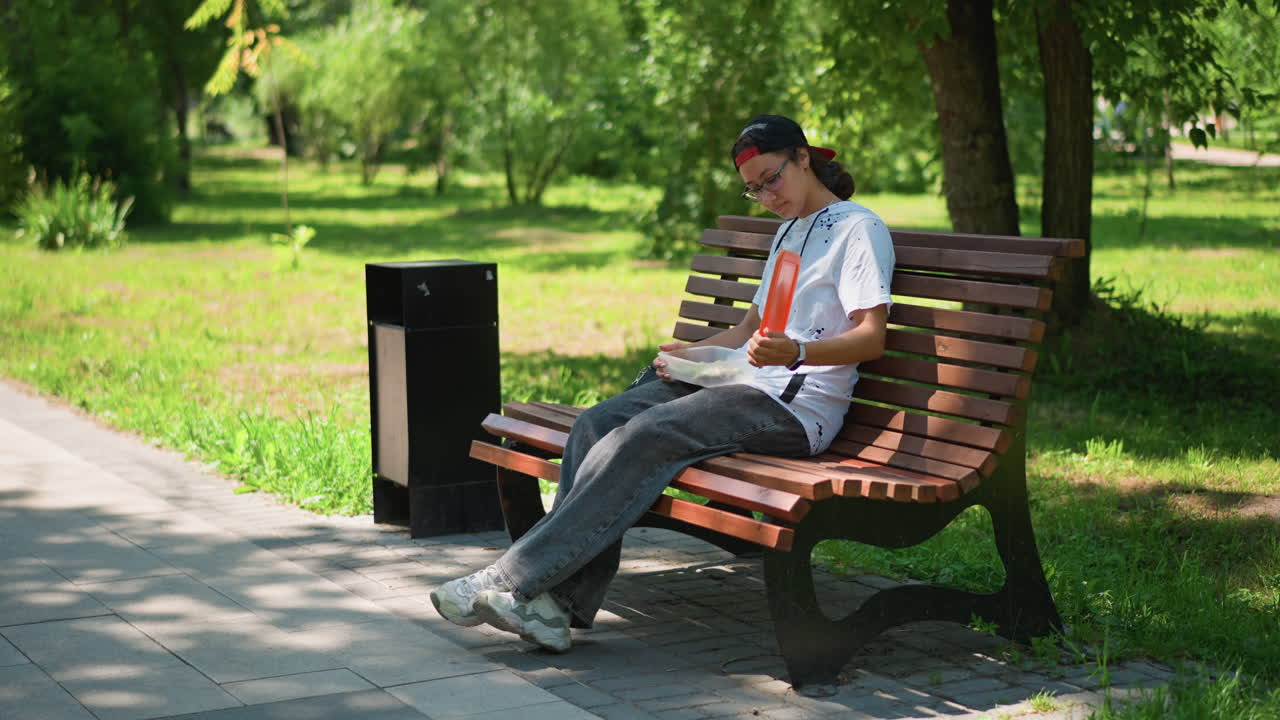 Hombre mirando el teléfono en un banco del parque cerca de una papelera, joven viajero con mochila consultando mensajes y ajustándose la gorra, mirando de reojo el camino arbolado, luz de sol cálida y sombra, momento relajado de un viajero, banco de madera