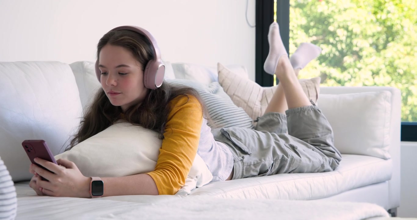 Ragazza adolescente con una maglia a maniche lunghe gialla sdraiata sul divano, il telefono riproduce audio, mentre allunga la mano per prendere le cuffie.