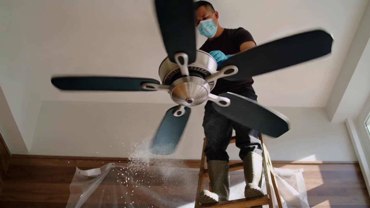 Uklízeč na žebříku drží točící se ventilátor, utírá prach na plast, má na sobě masku a rukavice, doma.