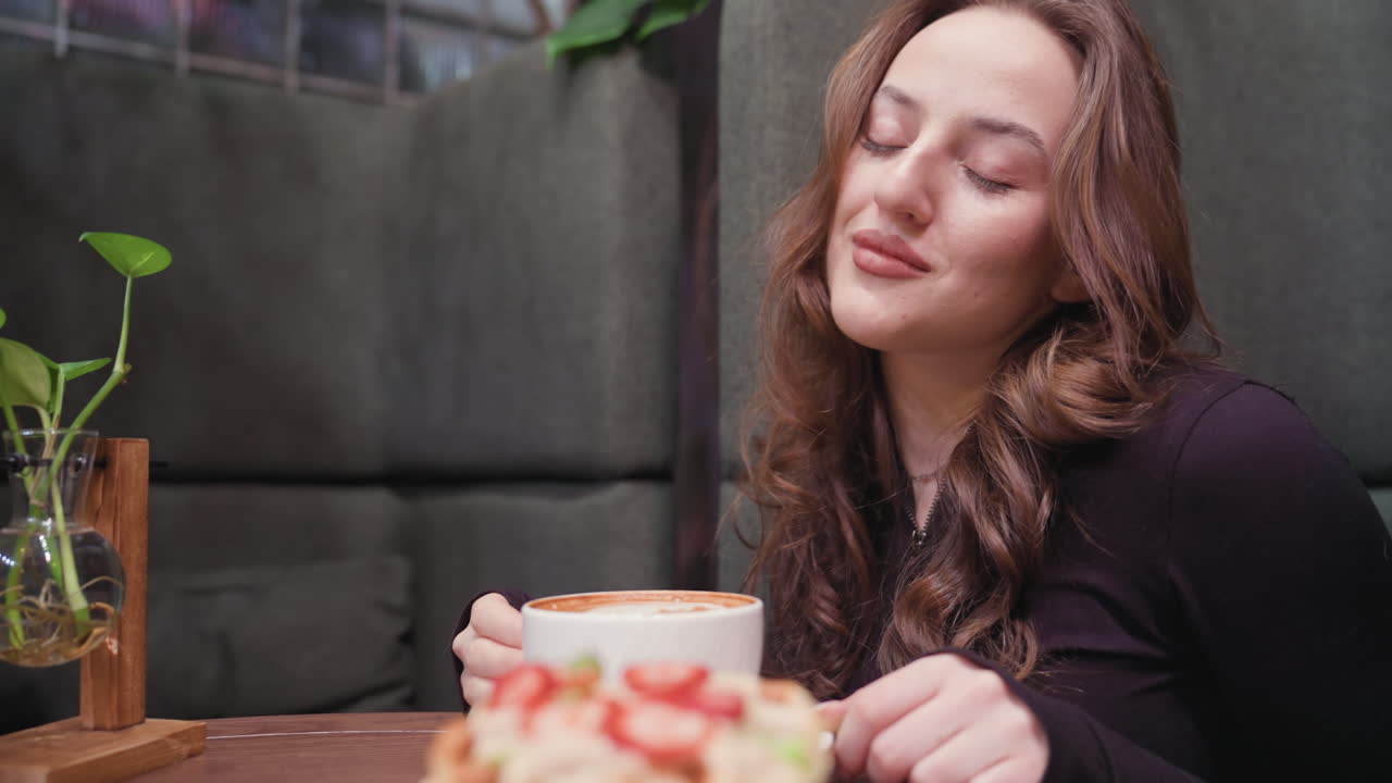 Young woman leans over white cup of latte, eyes closed, gently sniffing aroma. Decorative glass vase with green plant sits nearby on wooden table, creating cozy atmosphere with soft natural tones