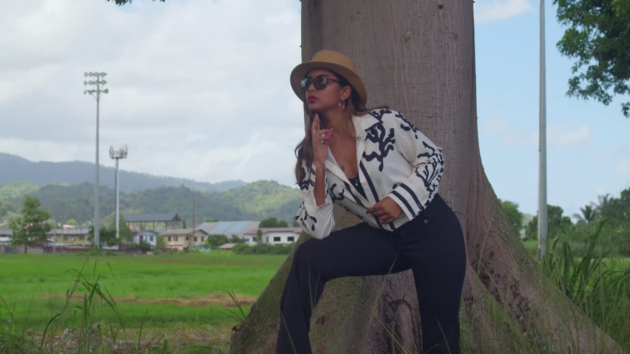 Una giovane donna con una camicia bianca abbottonata impeccabile e pantaloni eleganti blu navy siede su una panchina del parco con un cappello da sole, assorta in un romanzo sullo sfondo di montagne.