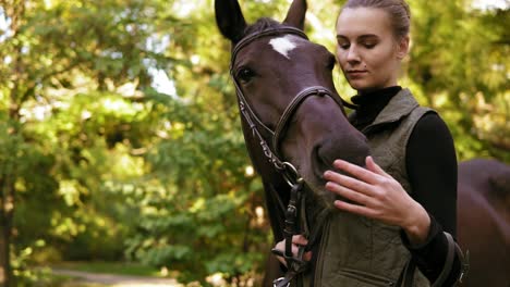 close up view of young beautiful woman petting brown horse while standing in the green field