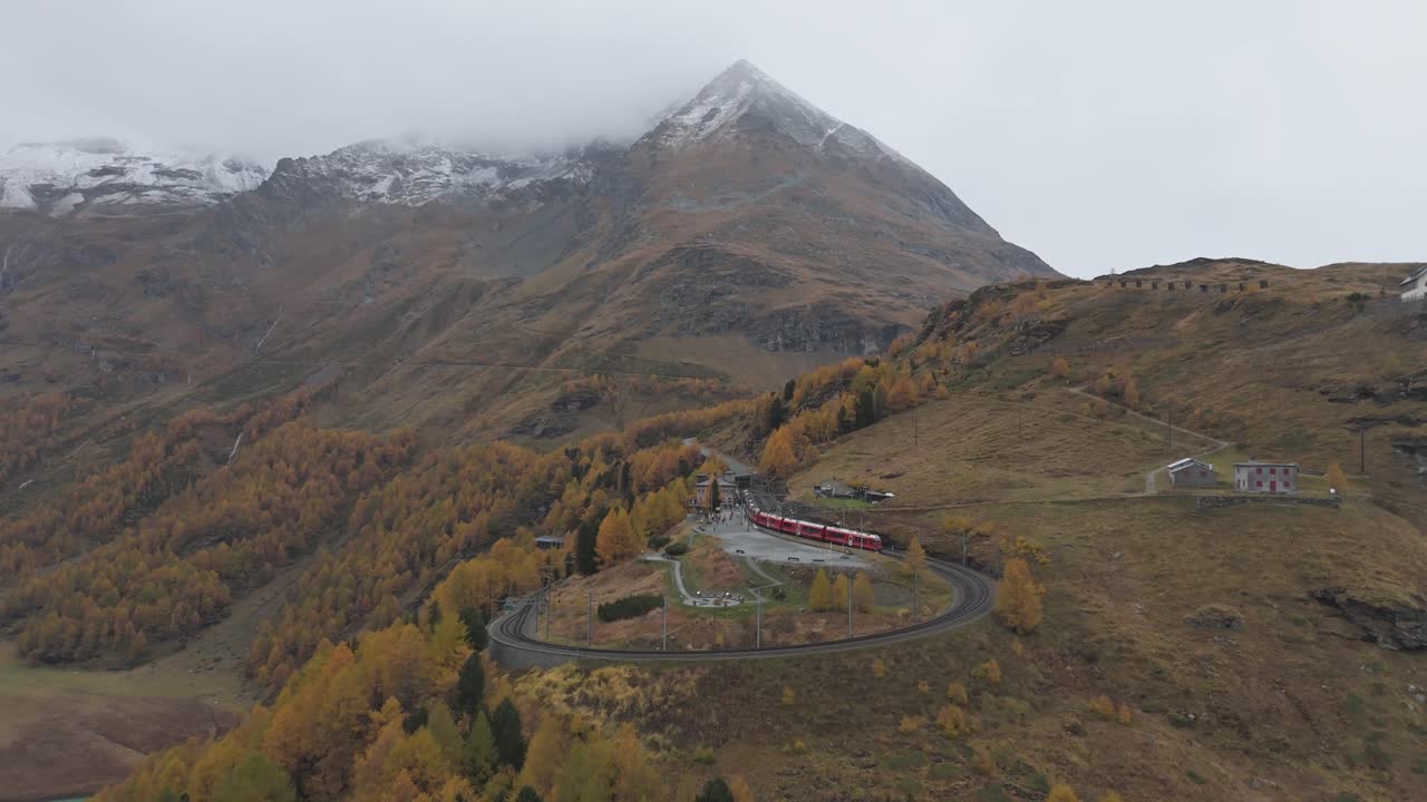 Red train of Bernina express is winding through autumnal landscape of swiss alps near on a cloudy day at the Alp Grüm stop, Graubünden, Switzerland, drobe revealing slow motion flying forward shot