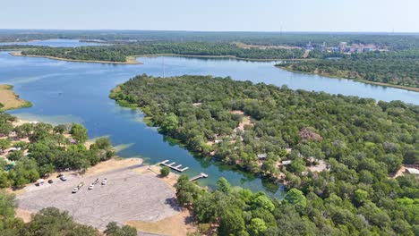 moving above a boat ramp parking lot, to the left above a pretty blue lake and recreational area that has shoreline and a campsite on the other side