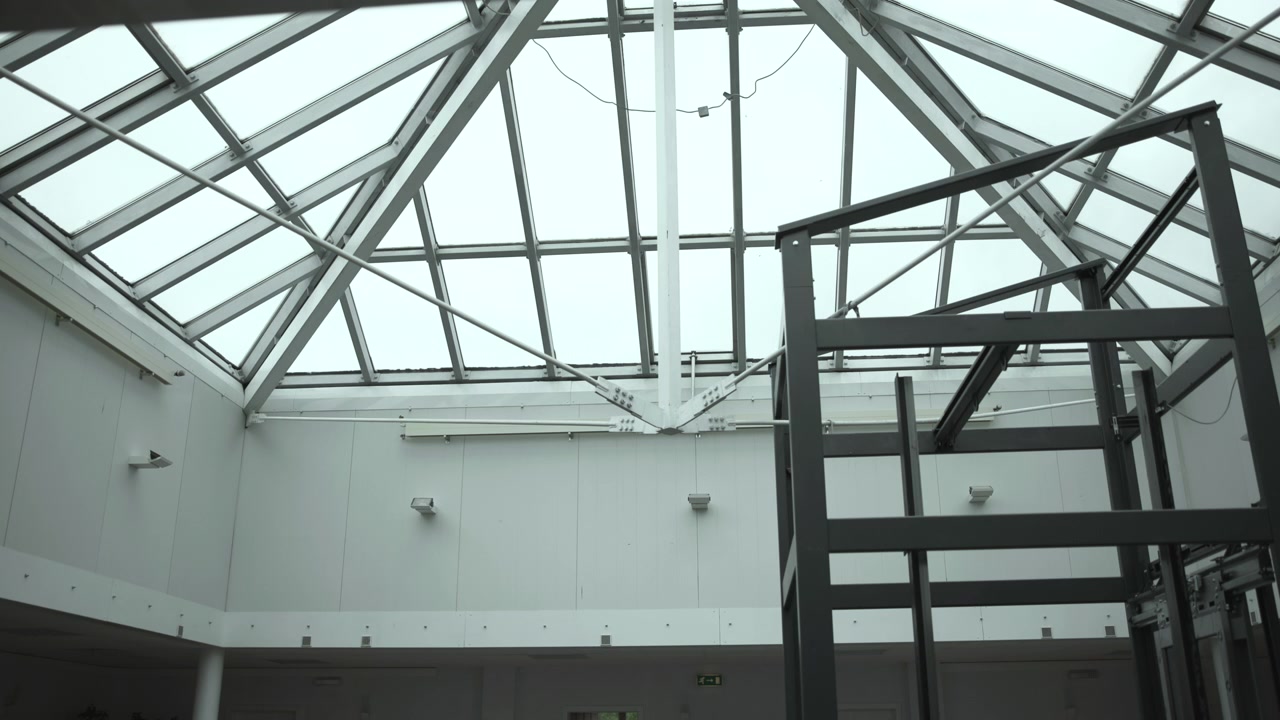 Low-angle interior view of industrial atrium showing pitched glass skylight, triangular steel trusses, diagonal braces, painted metal framework, cool diffuse daylight, and minimal functional