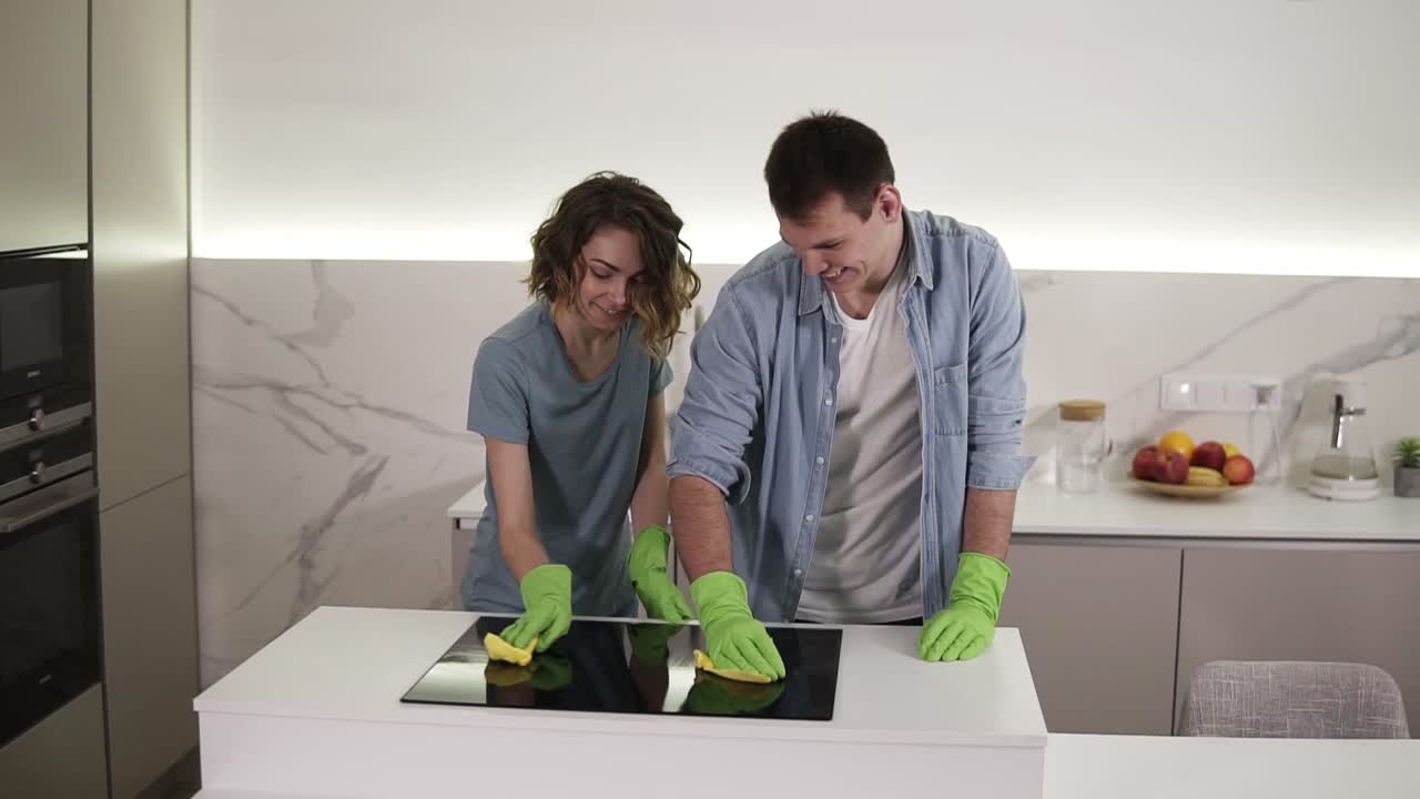 pareja con cuatro manos limpiando el panel de cocina de la cocina. joven y mujer limpiando la superficie perfecta de la estufa de cocina de cerámica negra usando guantes y escoba. divirtiéndose. cámara lenta