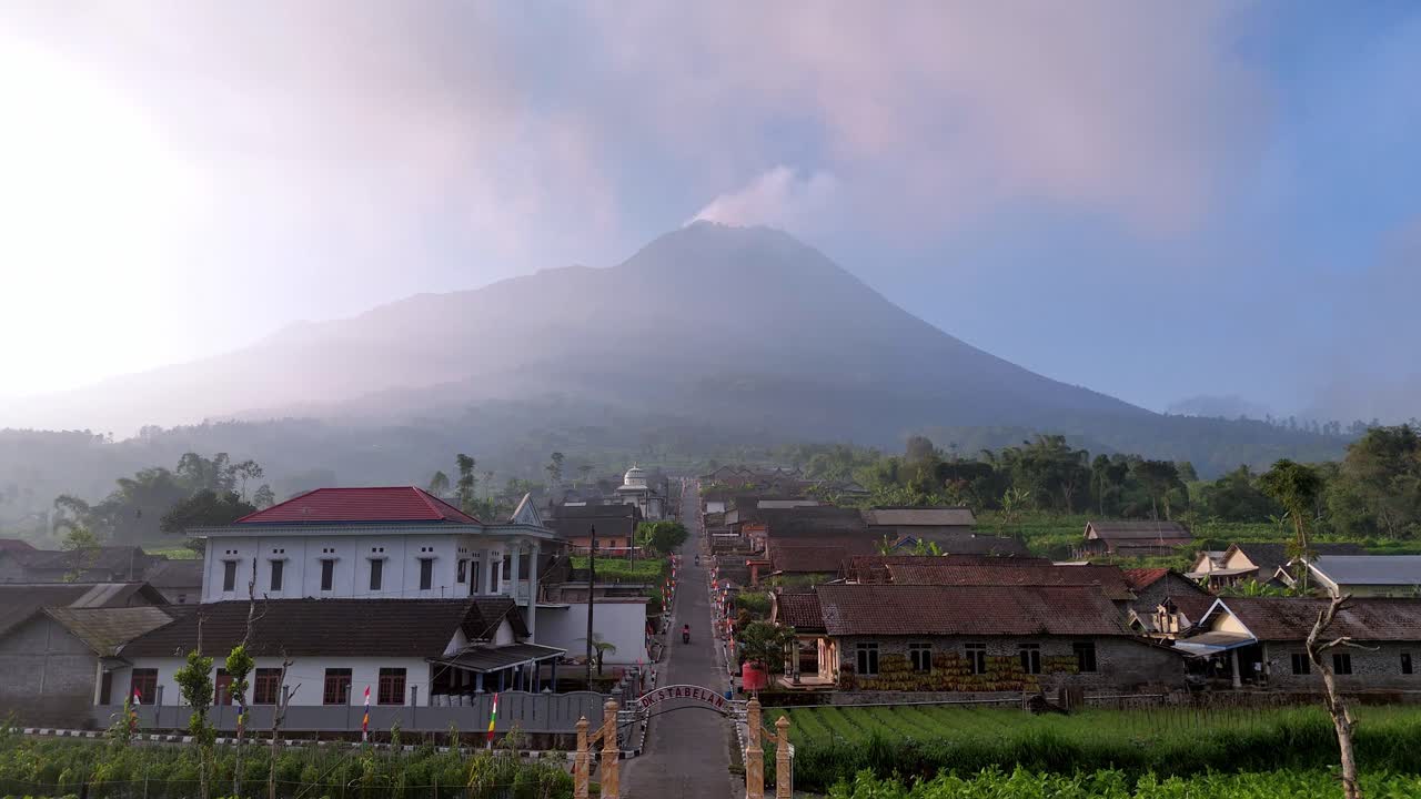 Aerial view of rural village with traditional houses and lush farmland, set against misty volcanic mountain in the morning light