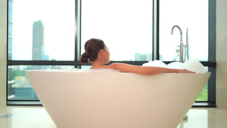 back view of sexy woman in luxury bathtub in front of windows with cityscape view