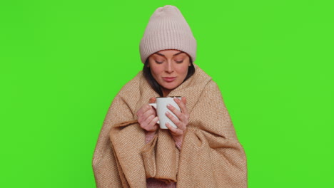 sick woman wear hat wrapped in plaid alone shivering from cold, drinking hot tea, try to warming up