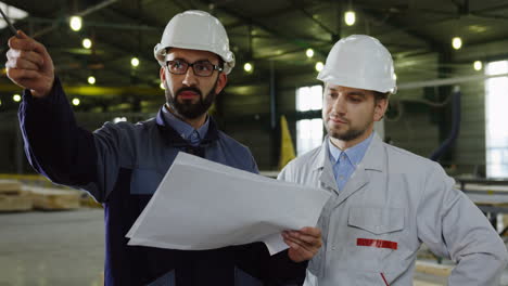 two workers wearing helmets talking while looking at blueprint in a big factory