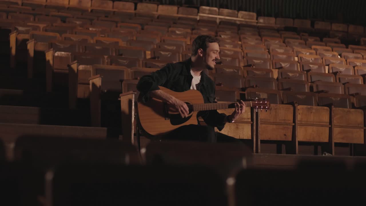un chico guapo toca una guitarra acústica en un cine abandonado. el músico canta una canción y acompaña en la guitarra