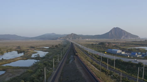 toma aérea de retroceso lento de un largo tren de carruajes de carbón completamente cargado, pasando por campos verdes, con una cresta montañosa a lo lejos, la carretera con camiones en movimiento en el lado derecho, al atardecer