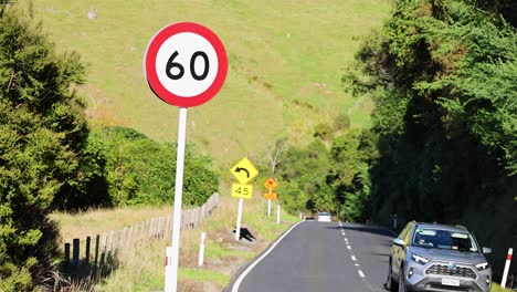 A car travels along a rural road in Akaroa, New Zealand, with clear skies and lush greenery. Bright daylight enhances visibility