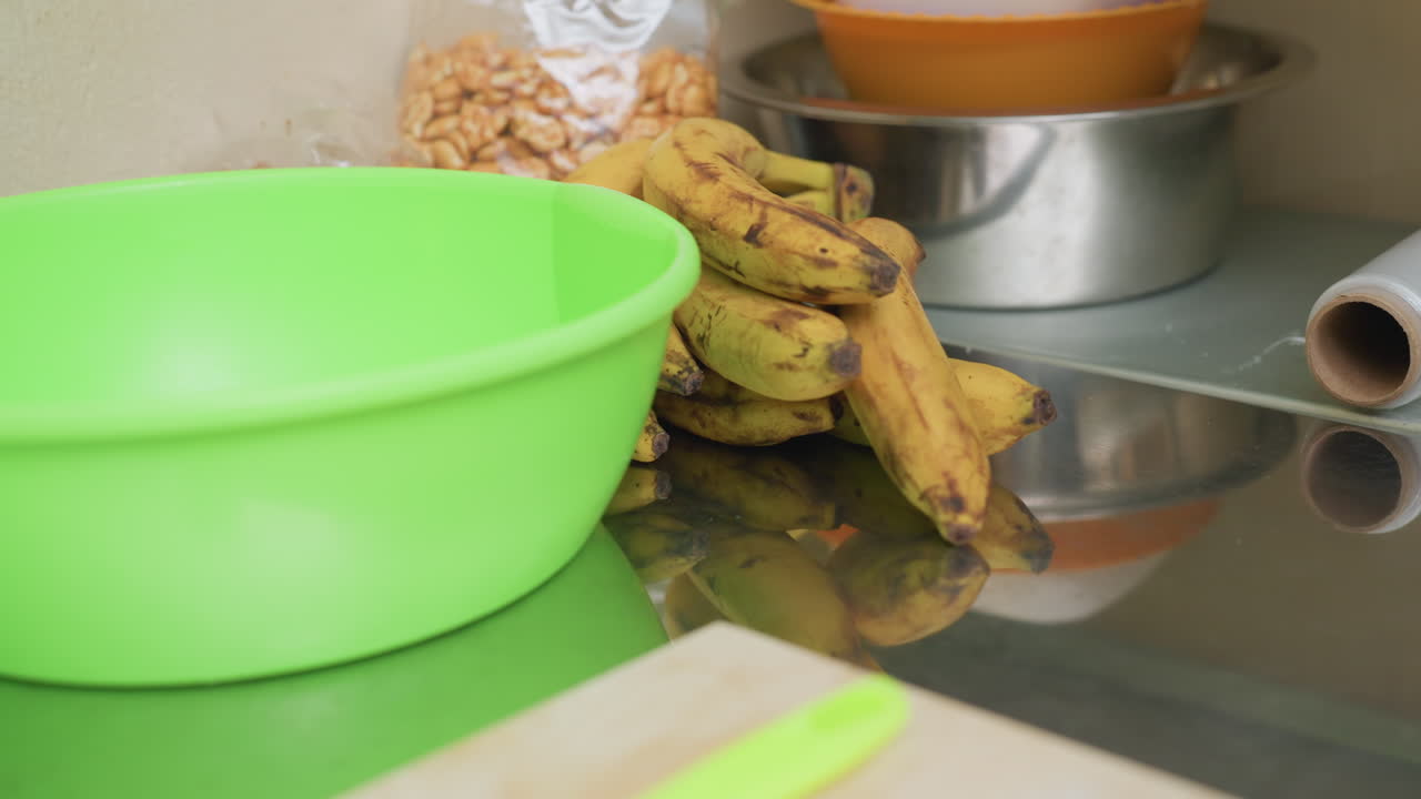 tigela verde na bancada com bananas, tigelas de aço inoxidável e saco de cereal ao fundo, rolo de papel e tábua de corte perto da espátula neon, close de um cacho de bananas maduras e uma mão alcançando a fruta, casual