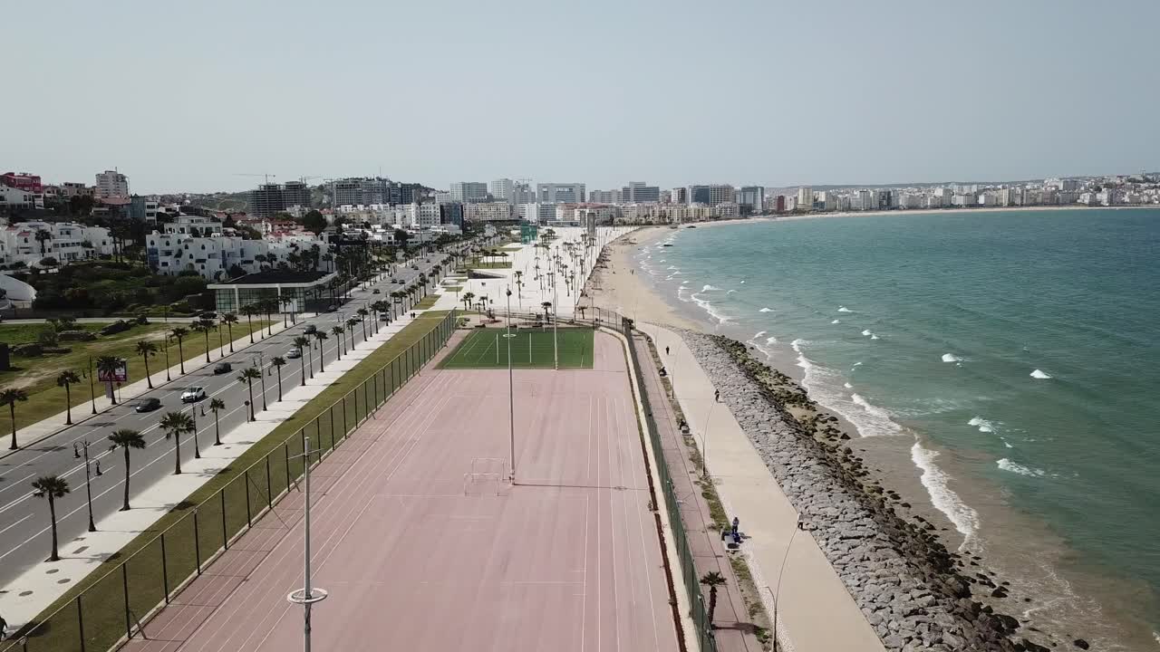 Vista aerea con drone di una passeggiata costiera con palme, strada lungomare e costa rocciosa lungo l'Oceano Atlantico, con lo skyline della città sullo sfondo in Marocco.
