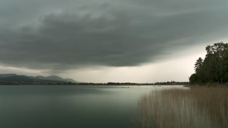 Grey-storm-clouds-pass-across-white-sky-over-lake-edge-with-wall-of-rain-in-wetzikon,-Switzerland,-wide-angle