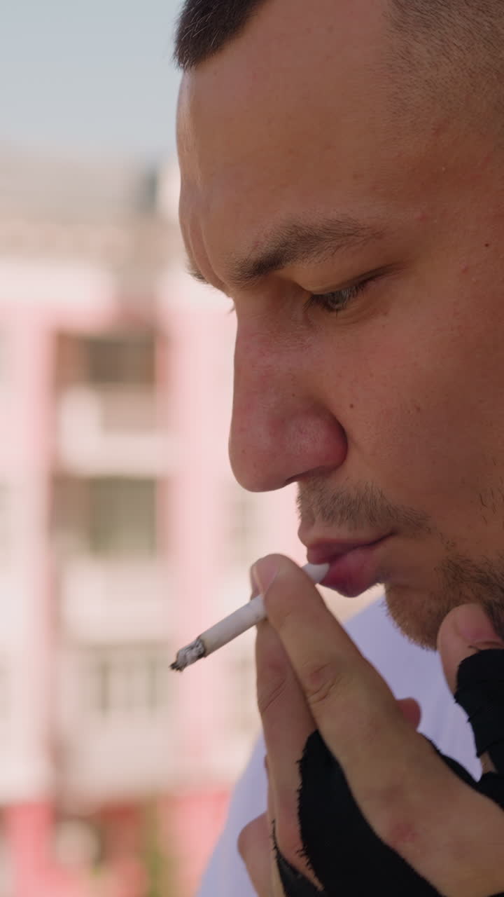 Attractive man in white gown with black glove smokes cigarette thinking on balcony of hotel room in morning close side view slow motion